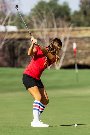 Female golfer about hit a short iron shot wearing knee-high PRO Compression socks in white with red and blue stripes.