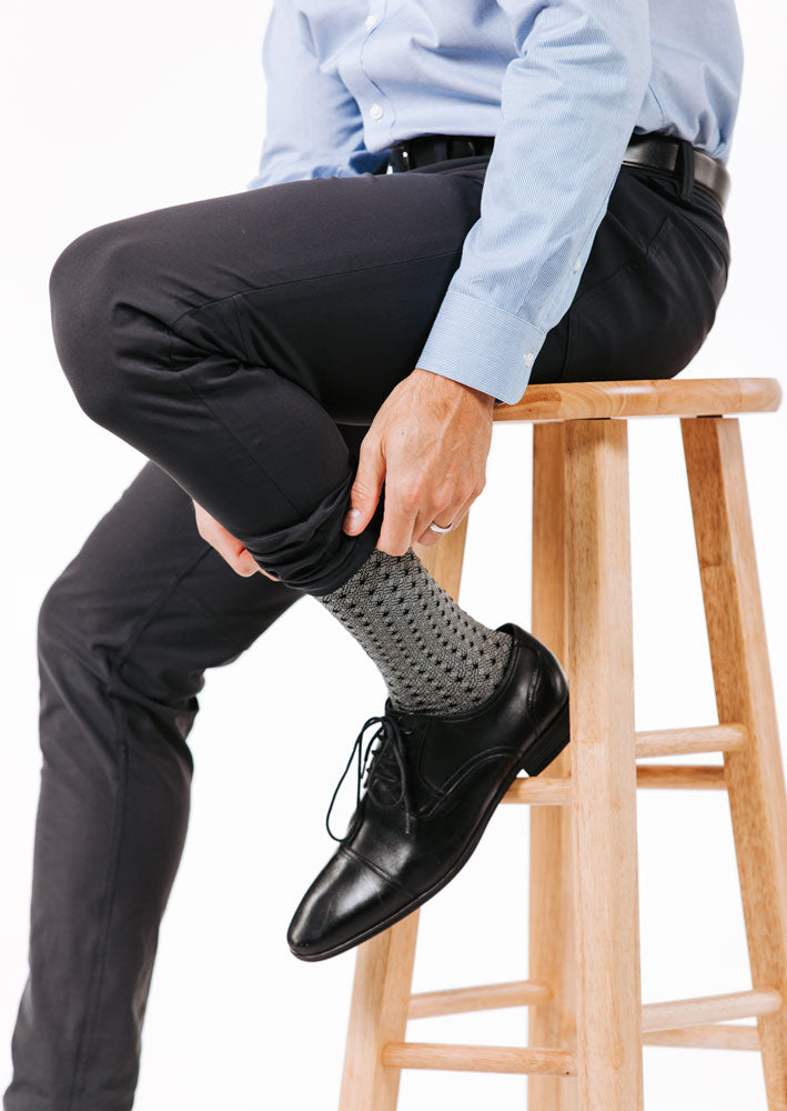 Person sitting on a wooden stool wearing black dress shoes, gray patterned compression socks, and black pants.
